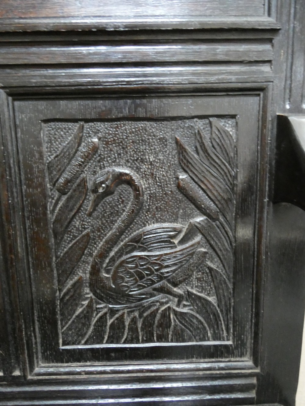 A dark carved oak high-back settle featuring relief panel carvings of swans and stags, turned arm supports, and a hinged box seat. View V