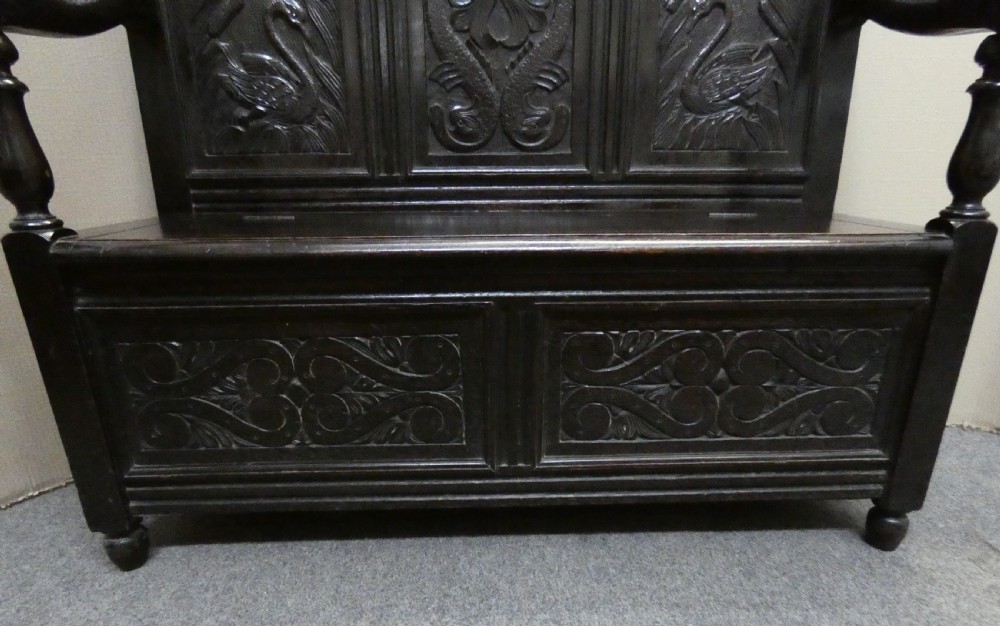 A dark carved oak high-back settle featuring relief panel carvings of swans and stags, turned arm supports, and a hinged box seat. View VII