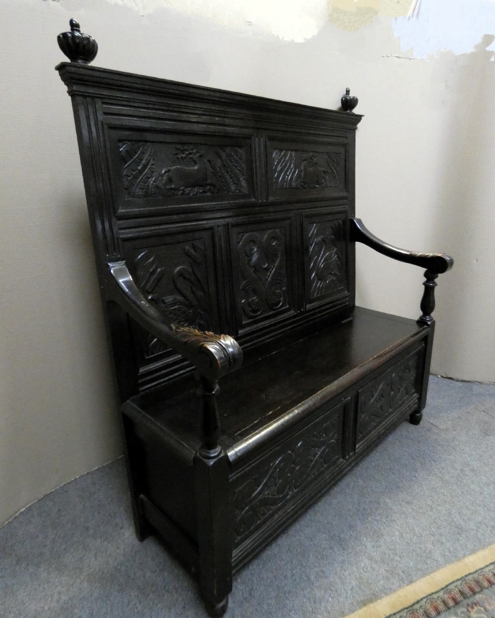 A dark carved oak high-back settle featuring relief panel carvings of swans and stags, turned arm supports, and a hinged box seat. View VIII
