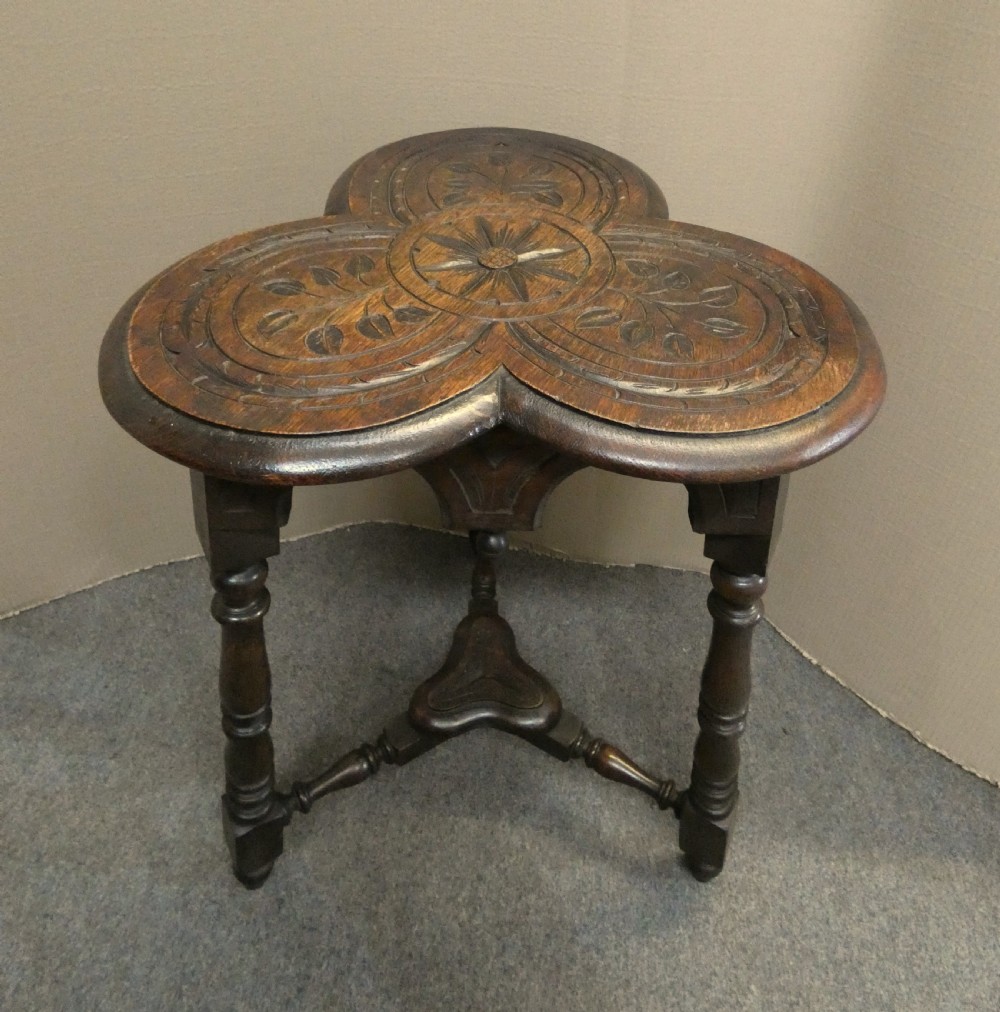 A dark oak Arts and Crafts occasional table featuring a three-lobed trefoil top carved with leaf motifs, set upon three turned legs joined by a lower stretcher.