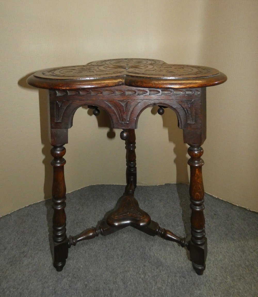 A dark oak Arts and Crafts occasional table featuring a three-lobed trefoil top carved with leaf motifs, set upon three turned legs joined by a lower stretcher. View II