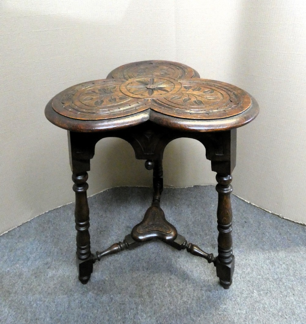 A dark oak Arts and Crafts occasional table featuring a three-lobed trefoil top carved with leaf motifs, set upon three turned legs joined by a lower stretcher. View III