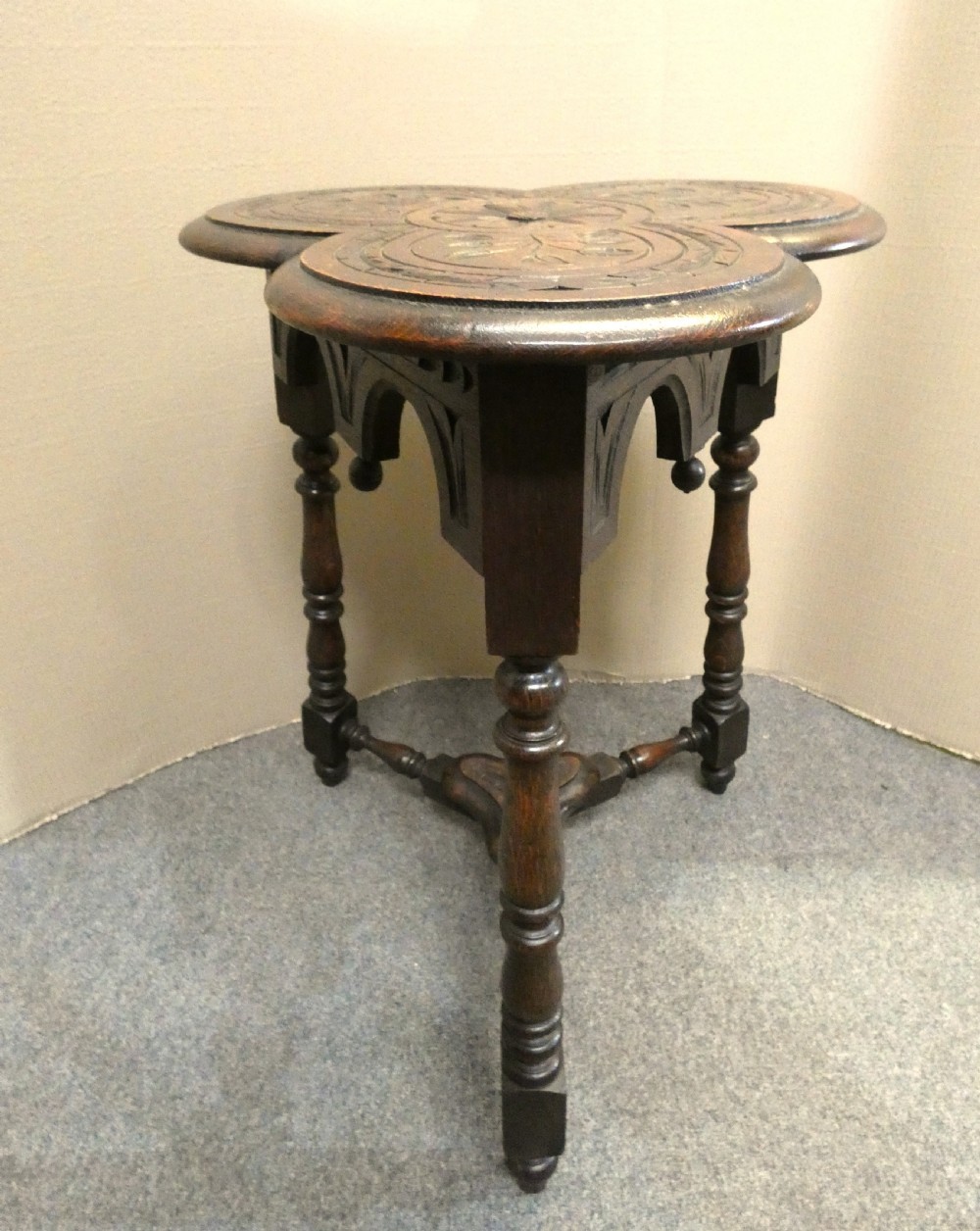 A dark oak Arts and Crafts occasional table featuring a three-lobed trefoil top carved with leaf motifs, set upon three turned legs joined by a lower stretcher. View V