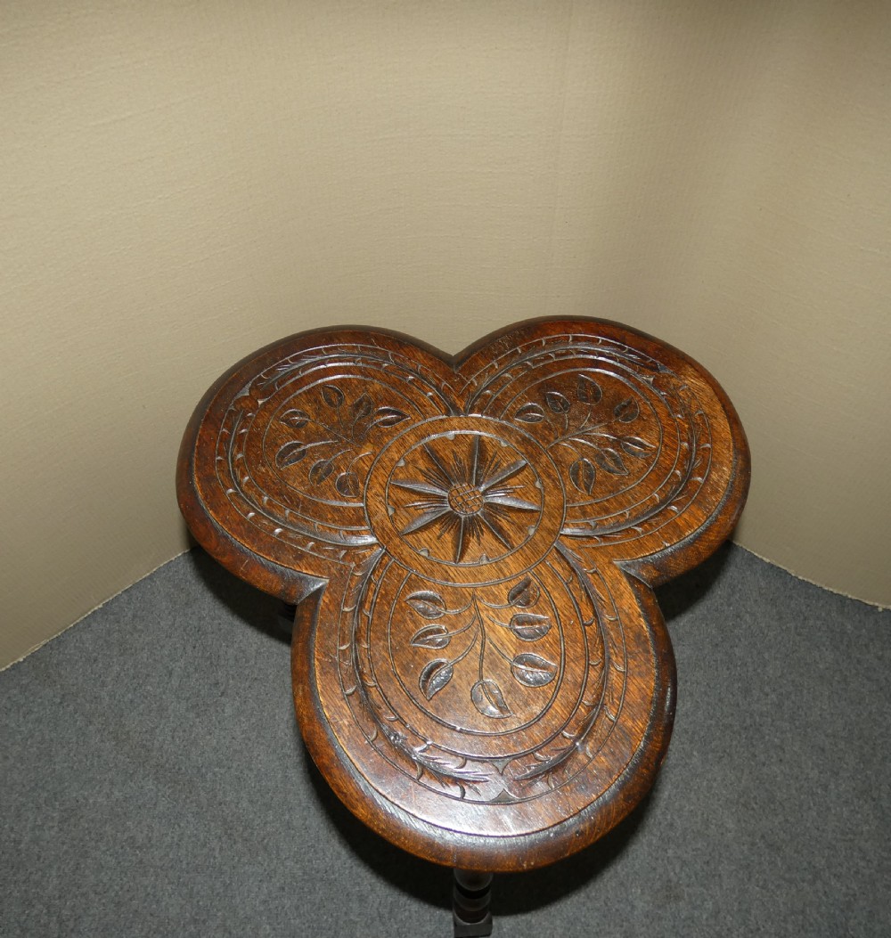 A dark oak Arts and Crafts occasional table featuring a three-lobed trefoil top carved with leaf motifs, set upon three turned legs joined by a lower stretcher. View VI