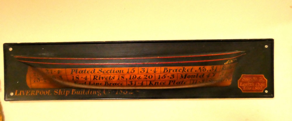A Victorian mounted half-hull ship model painted in terracotta and black with hand-written technical specifications on a black backboard.