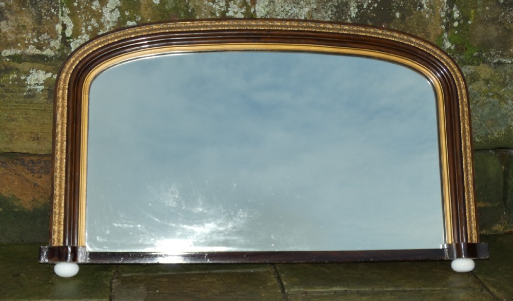A Victorian arched overmantel mirror featuring a dark timber frame, inner gilt bead moulding, and white porcelain corner feet.