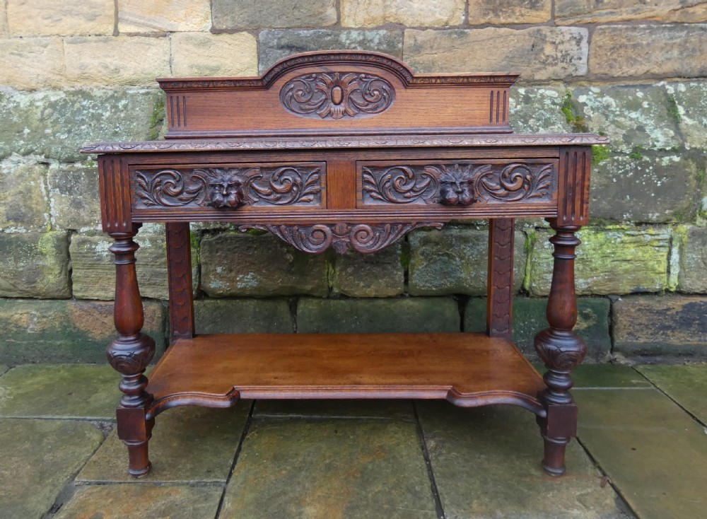 A dark carved oak hall table featuring a detailed relief gallery, two drawers with carved mask handles, turned legs, and a lower shelf.