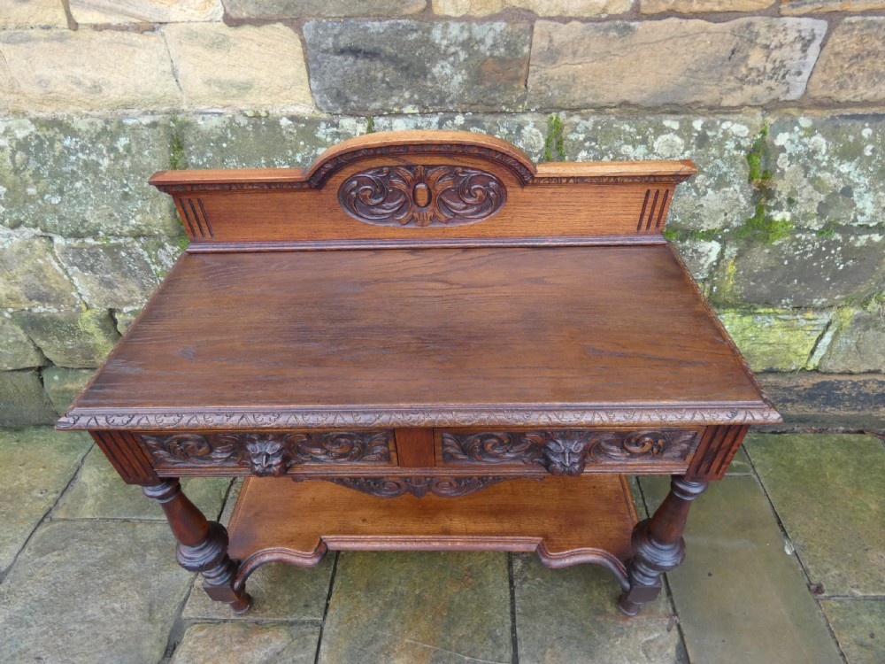 A dark carved oak hall table featuring a detailed relief gallery, two drawers with carved mask handles, turned legs, and a lower shelf. View III