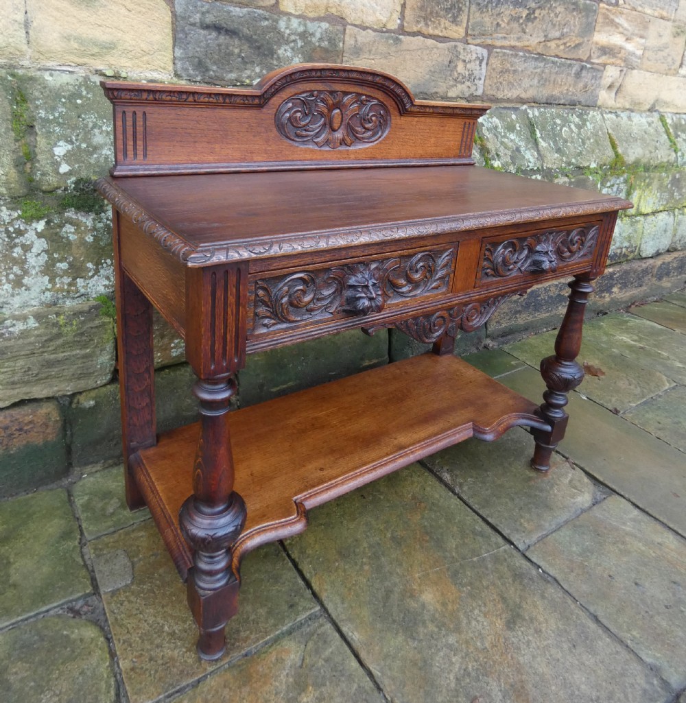 A dark carved oak hall table featuring a detailed relief gallery, two drawers with carved mask handles, turned legs, and a lower shelf. View V