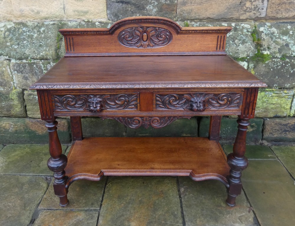 A dark carved oak hall table featuring a detailed relief gallery, two drawers with carved mask handles, turned legs, and a lower shelf. View VII