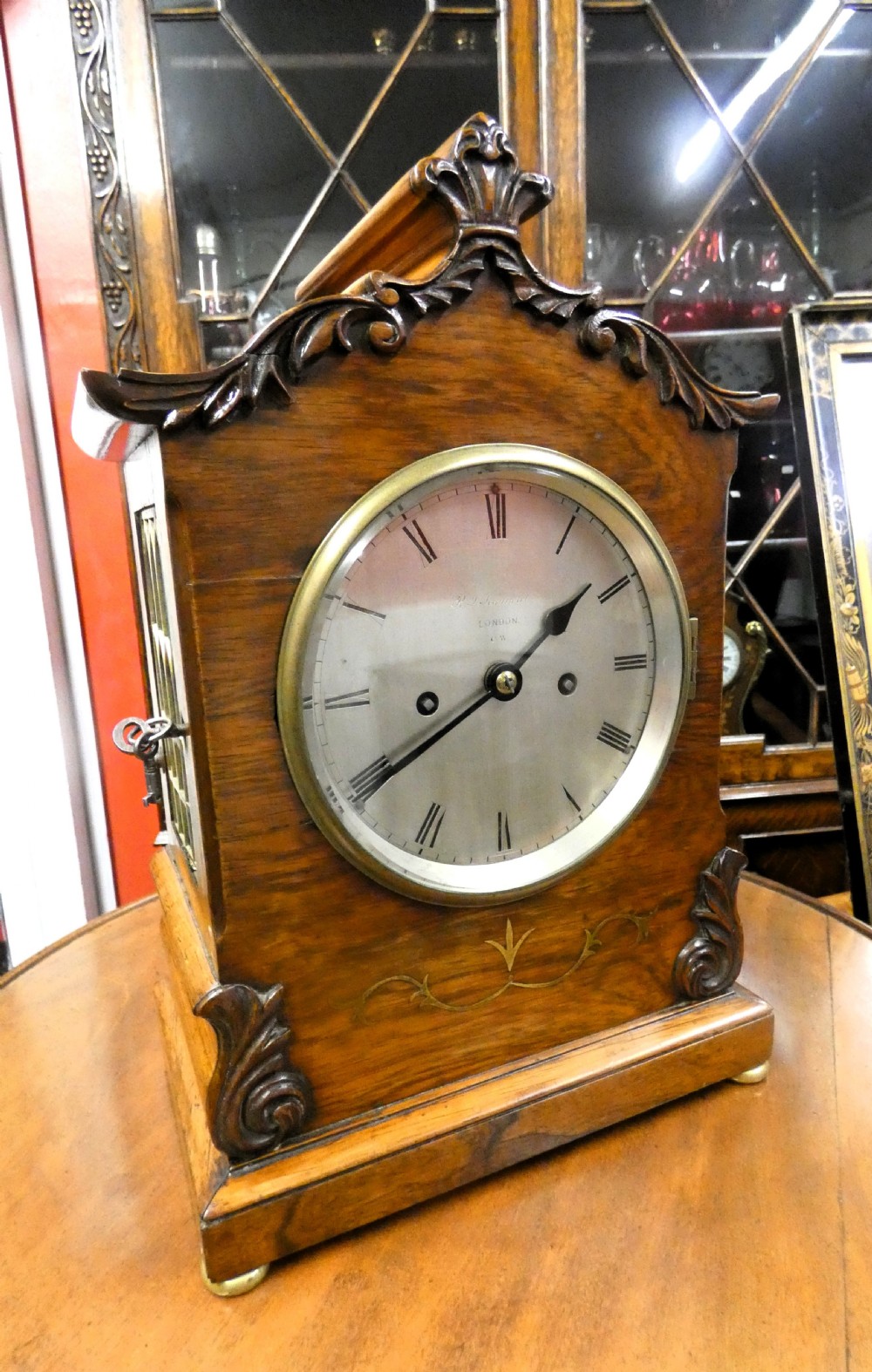 A William IV wooden bracket clock with carved foliate corner accents, brass scroll inlay, and a round dial displaying Roman numerals.