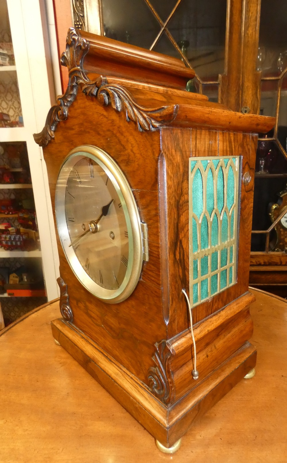 A William IV wooden bracket clock with carved foliate corner accents, brass scroll inlay, and a round dial displaying Roman numerals. View V