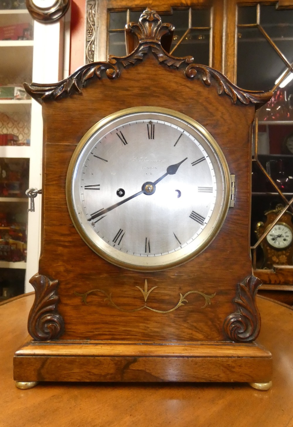 A William IV wooden bracket clock with carved foliate corner accents, brass scroll inlay, and a round dial displaying Roman numerals. View VI