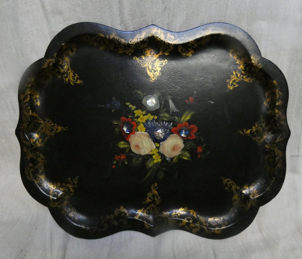 A black Victorian papier-mache serving tray featuring a scalloped rim, gilded scrollwork ornamentation, and a hand-painted floral centrepiece inlaid with mother-of-pearl.