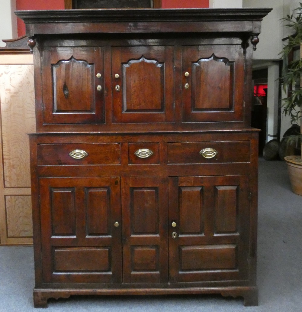 An antique dark oak Welsh deuddarn featuring a two-tiered design with arched fielded panels, brass handles, and carved drop pendants along the top canopy.