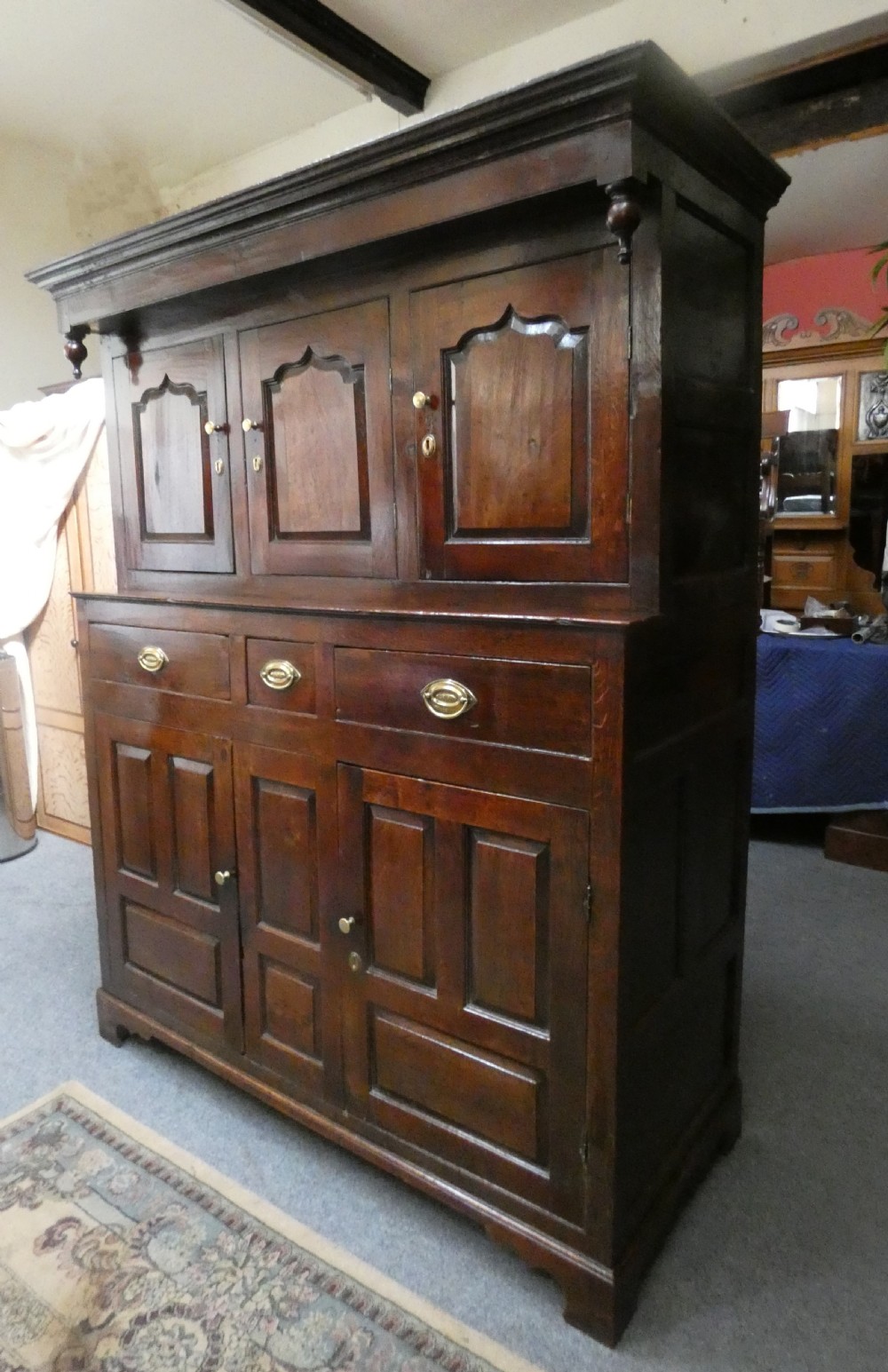 An antique dark oak Welsh deuddarn featuring a two-tiered design with arched fielded panels, brass handles, and carved drop pendants along the top canopy. View II