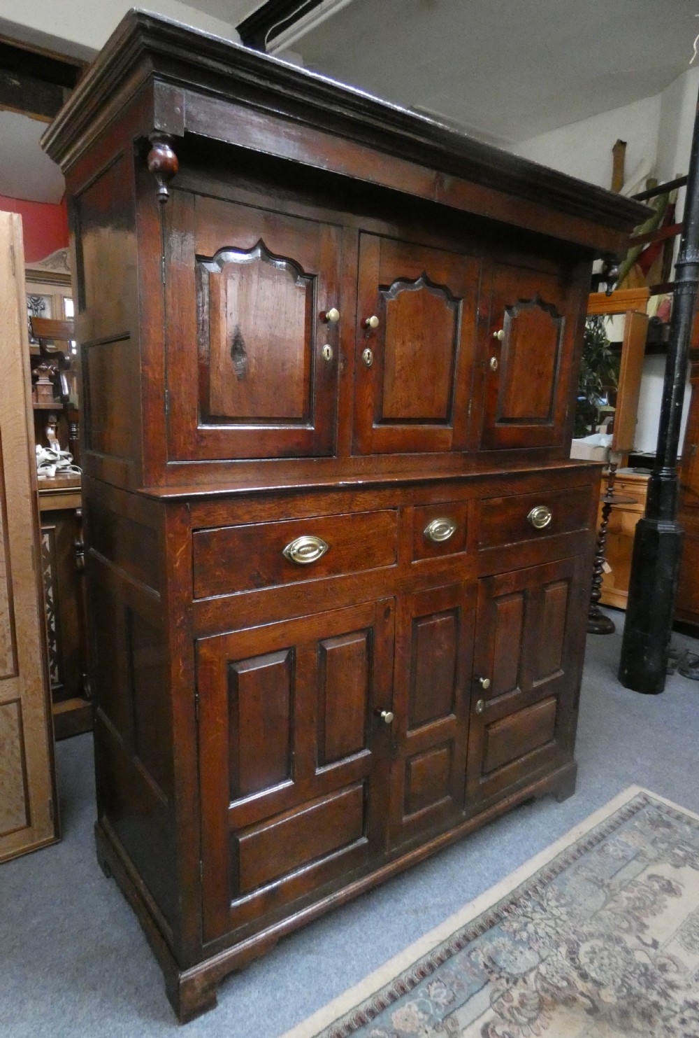 An antique dark oak Welsh deuddarn featuring a two-tiered design with arched fielded panels, brass handles, and carved drop pendants along the top canopy. View III