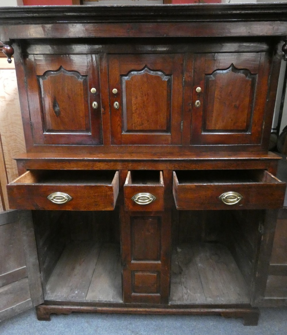 An antique dark oak Welsh deuddarn featuring a two-tiered design with arched fielded panels, brass handles, and carved drop pendants along the top canopy. View VII