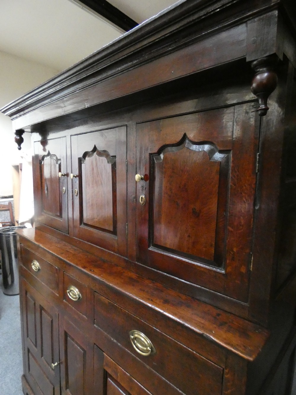 An antique dark oak Welsh deuddarn featuring a two-tiered design with arched fielded panels, brass handles, and carved drop pendants along the top canopy. View IX