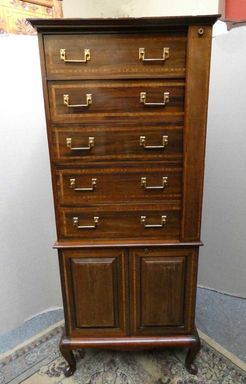 A tall antique mahogany collectors cabinet featuring five shallow upper drawers with brass handles, a hinged side locking bar, and a two-door lower cupboard resting on cabriole legs.