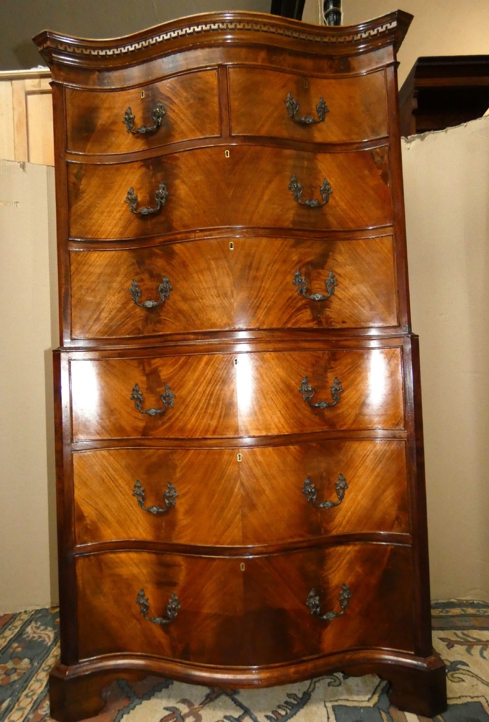 A Georgian flame mahogany chest-on-chest featuring a serpentine front, dentil moulding along the upper cornice, dark brass handles, and bracket feet. View V