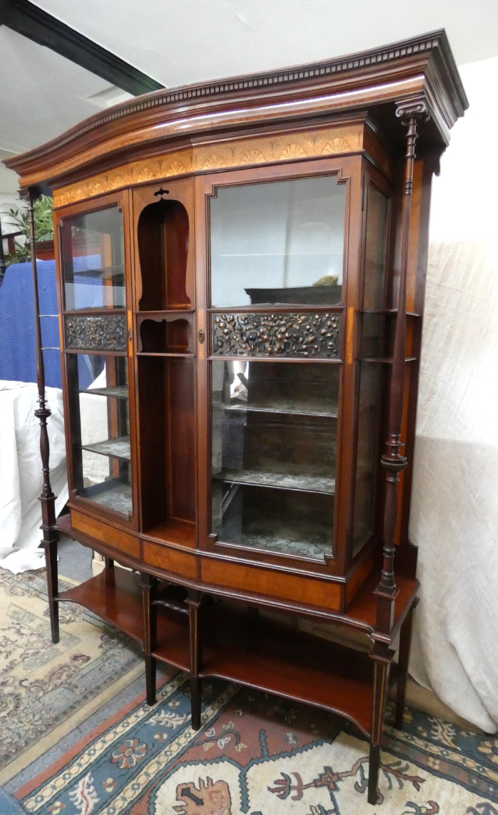 An antique mahogany display cabinet featuring inlaid satinwood decoration, carved floral door panels, glass display sections, and slender corner supports.