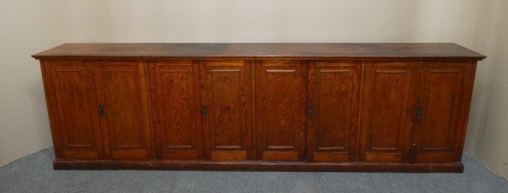 A long, low antique pitch pine cupboard featuring eight panelled doors with central keyhole escutcheons resting on a solid plinth base.