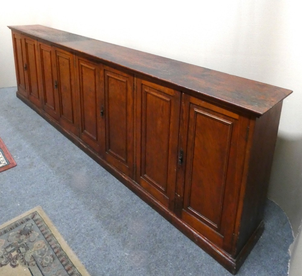 A long, low antique pitch pine cupboard featuring eight panelled doors with central keyhole escutcheons resting on a solid plinth base. View II