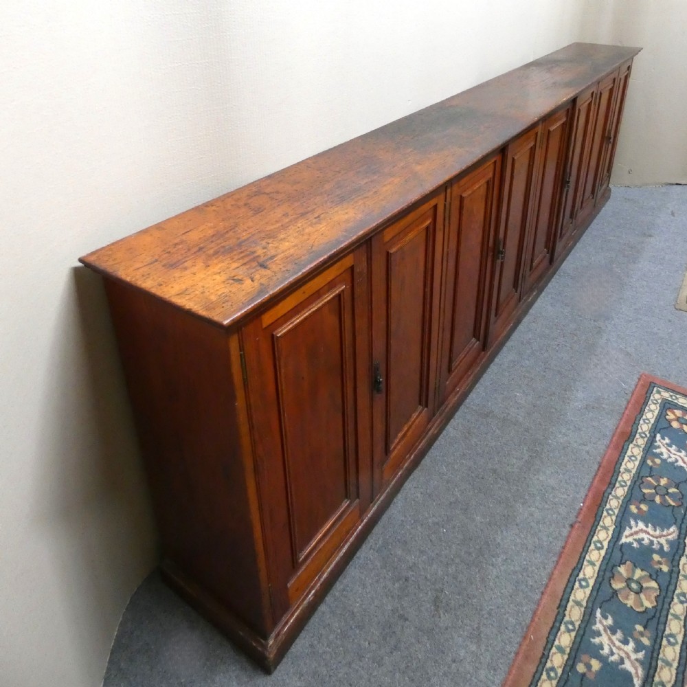A long, low antique pitch pine cupboard featuring eight panelled doors with central keyhole escutcheons resting on a solid plinth base. View III