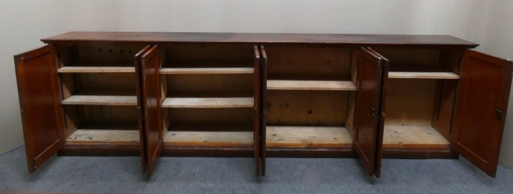 A long, low antique pitch pine cupboard featuring eight panelled doors with central keyhole escutcheons resting on a solid plinth base. View VII