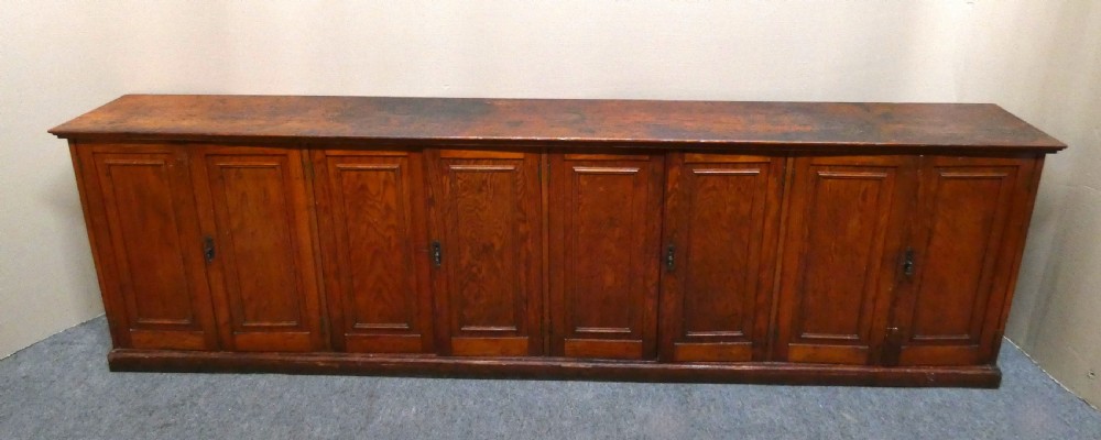 A long, low antique pitch pine cupboard featuring eight panelled doors with central keyhole escutcheons resting on a solid plinth base. View VIII