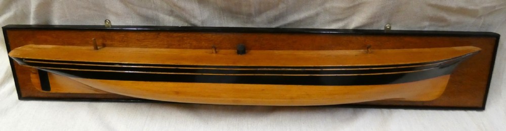 A carved wooden half-hull boat model with painted black accents, mounted on a dark wooden plaque.