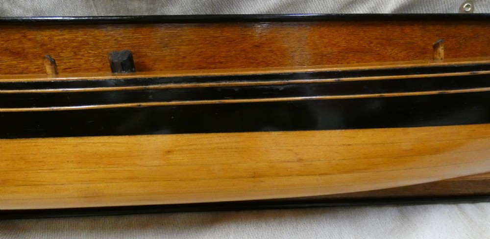 A carved wooden half-hull boat model with painted black accents, mounted on a dark wooden plaque. View VI
