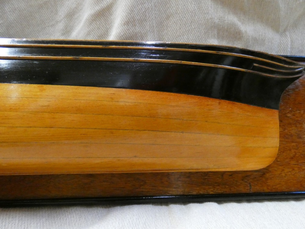 A carved wooden half-hull boat model with painted black accents, mounted on a dark wooden plaque. View VII