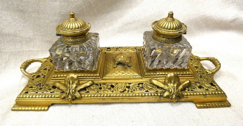 An ornate Victorian cast brass inkstand featuring twin faceted glass inkwells, a central tortoise motif, insect pen rests, and pierced scrollwork.