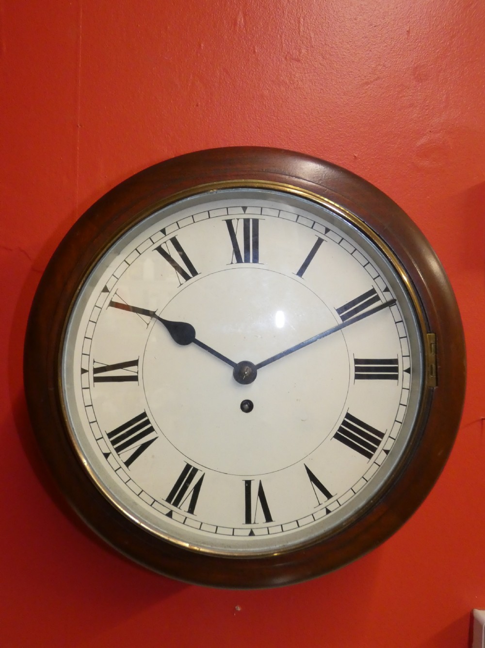 An antique circular wall clock featuring a turned mahogany surround, white painted dial with black Roman numerals, and a brass glass bezel. View III