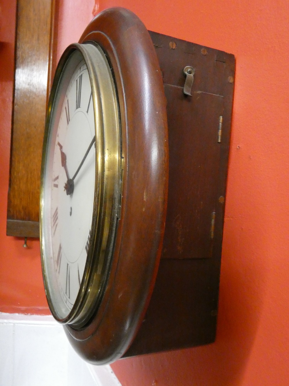 An antique circular wall clock featuring a turned mahogany surround, white painted dial with black Roman numerals, and a brass glass bezel. View V