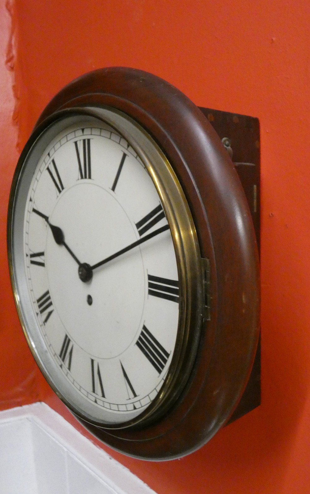 An antique circular wall clock featuring a turned mahogany surround, white painted dial with black Roman numerals, and a brass glass bezel. View VI