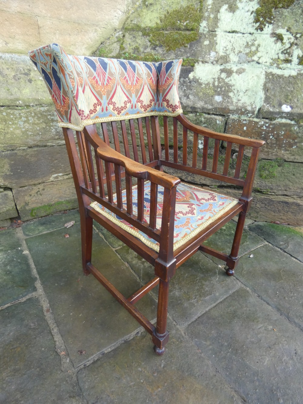 An Arts and Crafts wooden armchair featuring a slatted backrest, shaped armrests, turned front legs, and colourful patterned fabric upholstery. View II