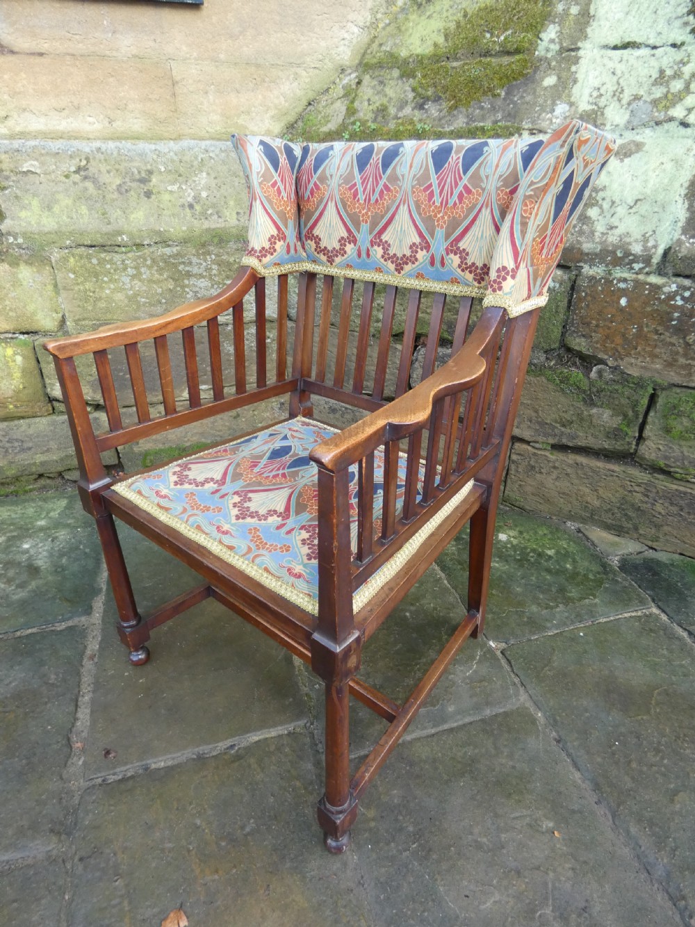 An Arts and Crafts wooden armchair featuring a slatted backrest, shaped armrests, turned front legs, and colourful patterned fabric upholstery. View III