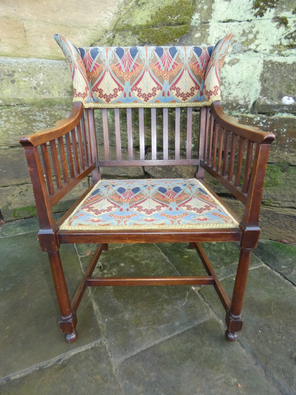 An Arts and Crafts wooden armchair featuring a slatted backrest, shaped armrests, turned front legs, and colourful patterned fabric upholstery. View V
