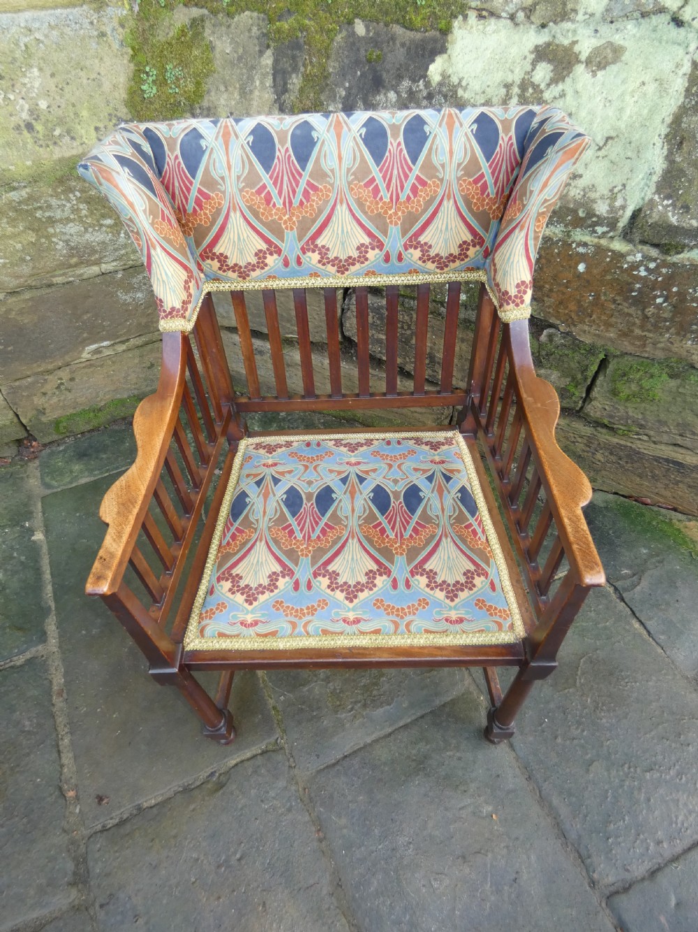 An Arts and Crafts wooden armchair featuring a slatted backrest, shaped armrests, turned front legs, and colourful patterned fabric upholstery. View VI