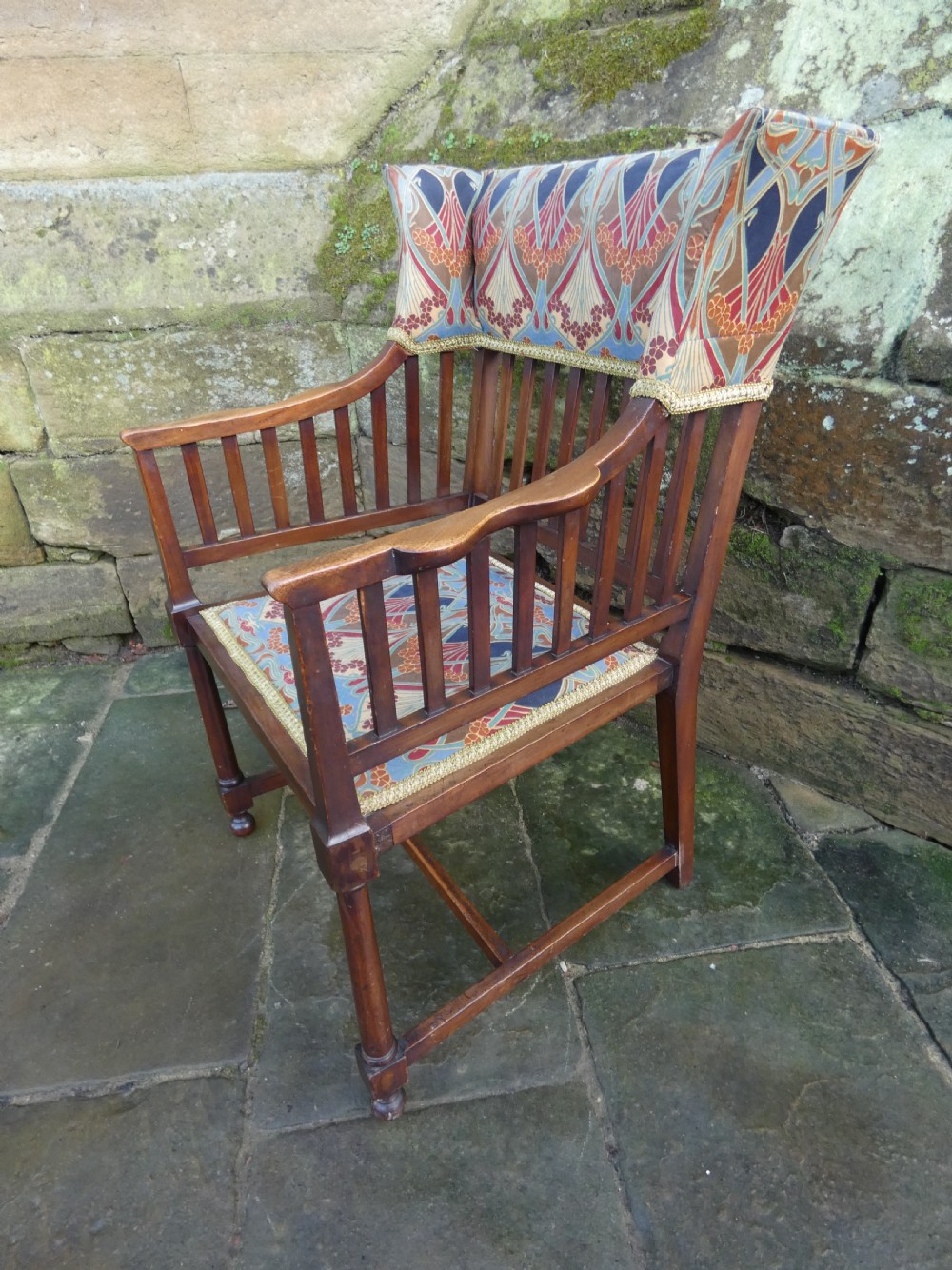 An Arts and Crafts wooden armchair featuring a slatted backrest, shaped armrests, turned front legs, and colourful patterned fabric upholstery. View VII
