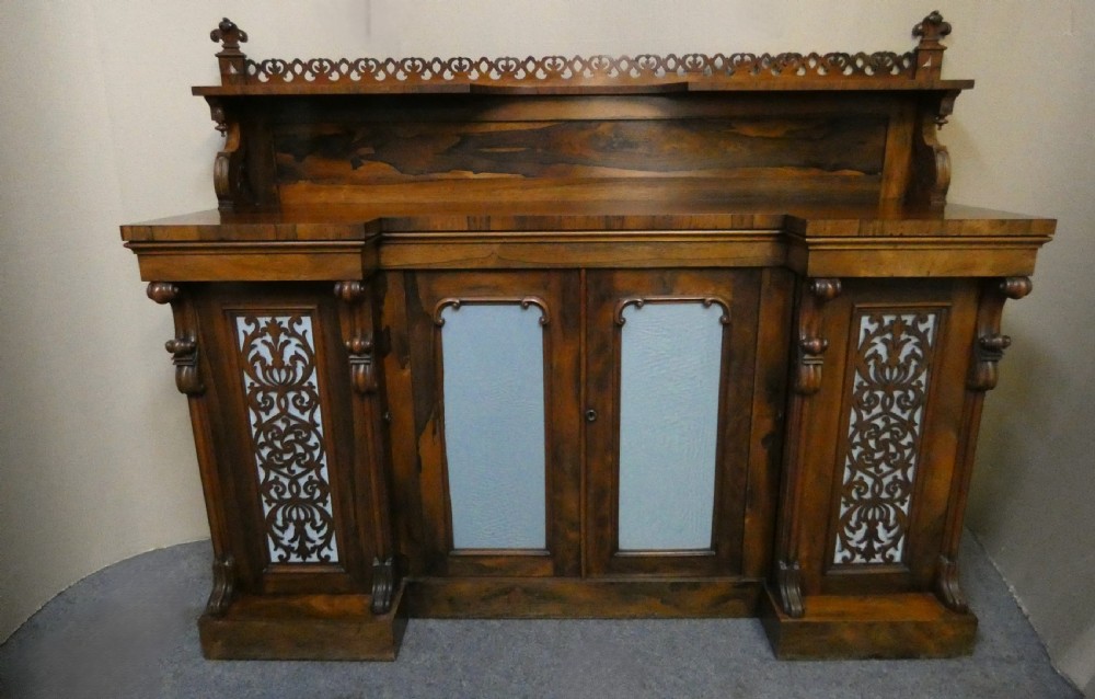 A Victorian rosewood breakfront chiffonier featuring a upper gallery shelf, central panelled cupboard doors, and outer doors with intricate pierced fretwork over light blue fabric backing.