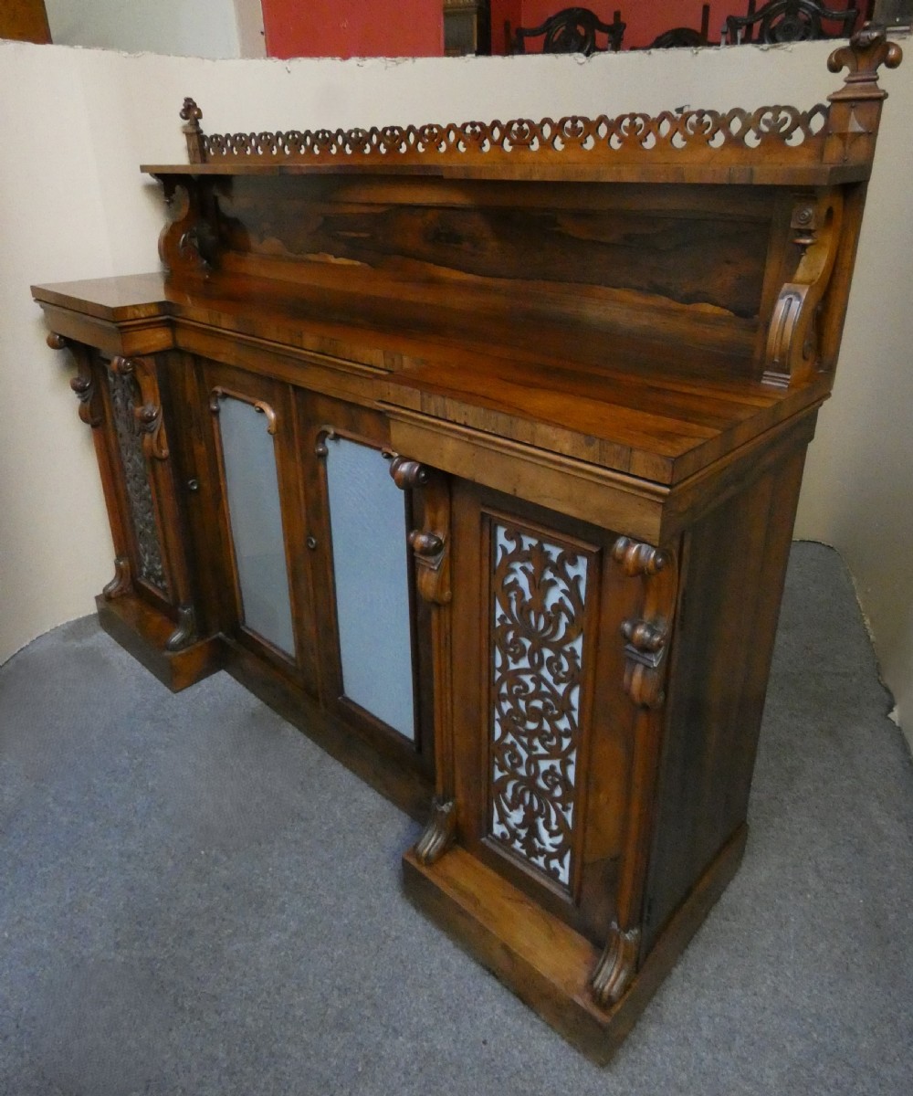 A Victorian rosewood breakfront chiffonier featuring a upper gallery shelf, central panelled cupboard doors, and outer doors with intricate pierced fretwork over light blue fabric backing. View II