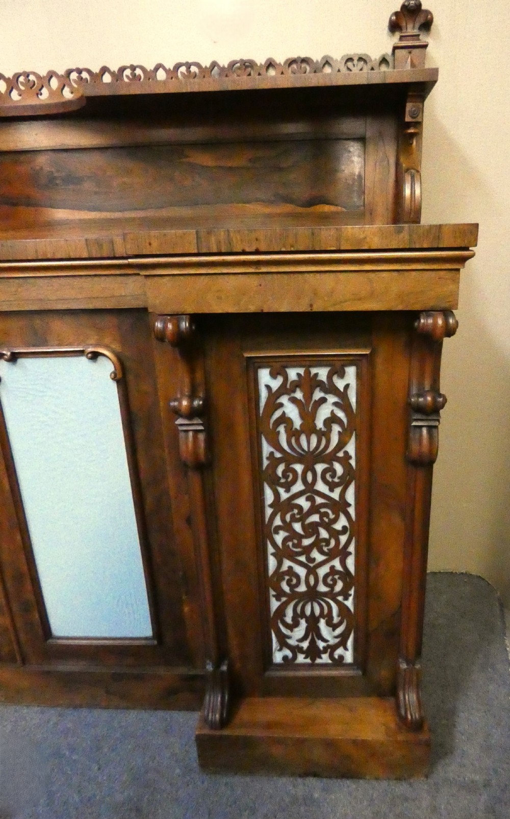 A Victorian rosewood breakfront chiffonier featuring a upper gallery shelf, central panelled cupboard doors, and outer doors with intricate pierced fretwork over light blue fabric backing. View III