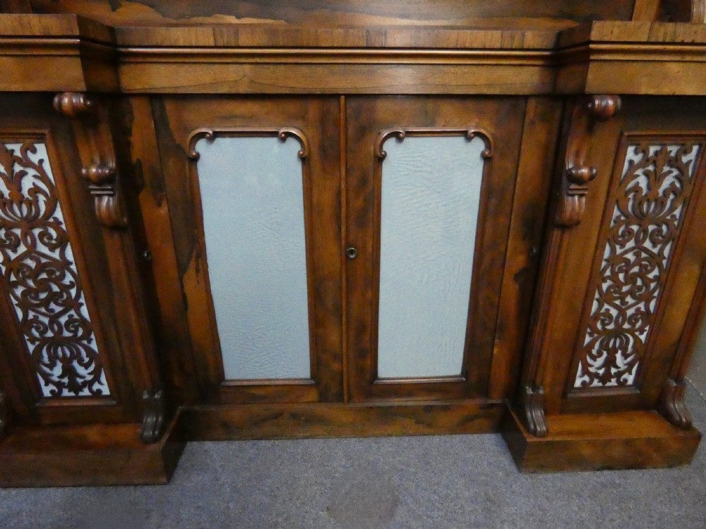 A Victorian rosewood breakfront chiffonier featuring a upper gallery shelf, central panelled cupboard doors, and outer doors with intricate pierced fretwork over light blue fabric backing. View VIII
