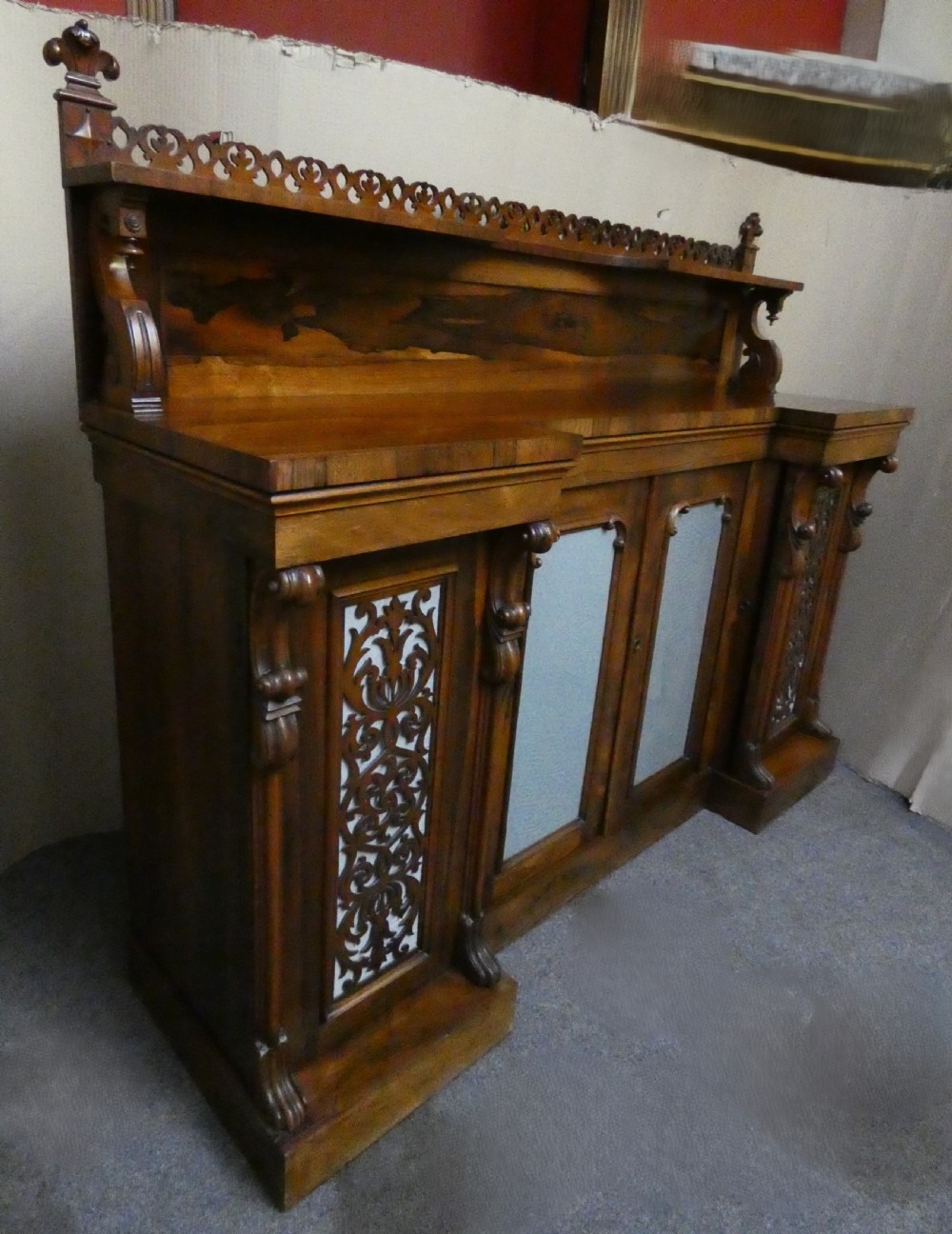 A Victorian rosewood breakfront chiffonier featuring a upper gallery shelf, central panelled cupboard doors, and outer doors with intricate pierced fretwork over light blue fabric backing. View X