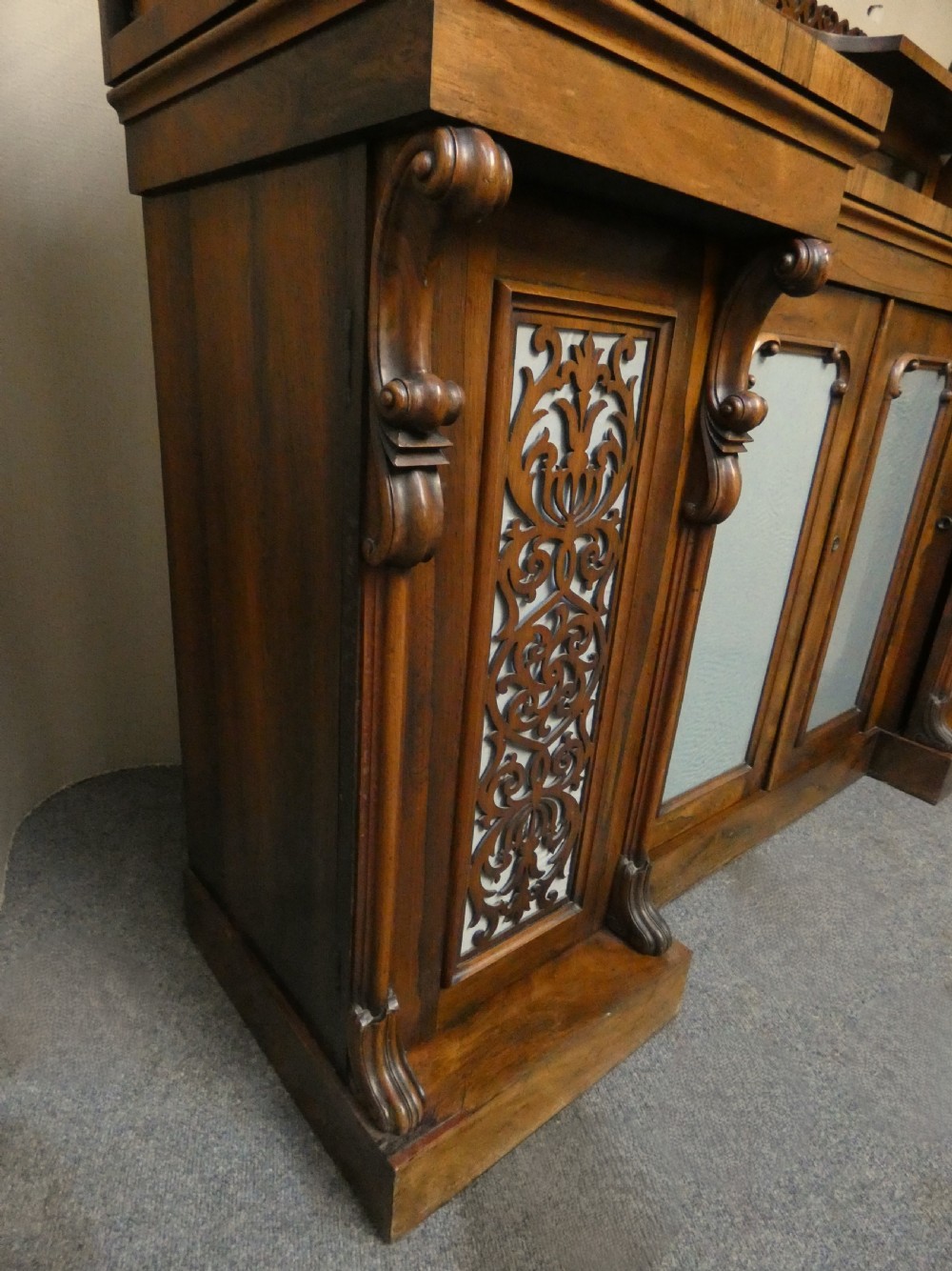 A Victorian rosewood breakfront chiffonier featuring a upper gallery shelf, central panelled cupboard doors, and outer doors with intricate pierced fretwork over light blue fabric backing. View XI