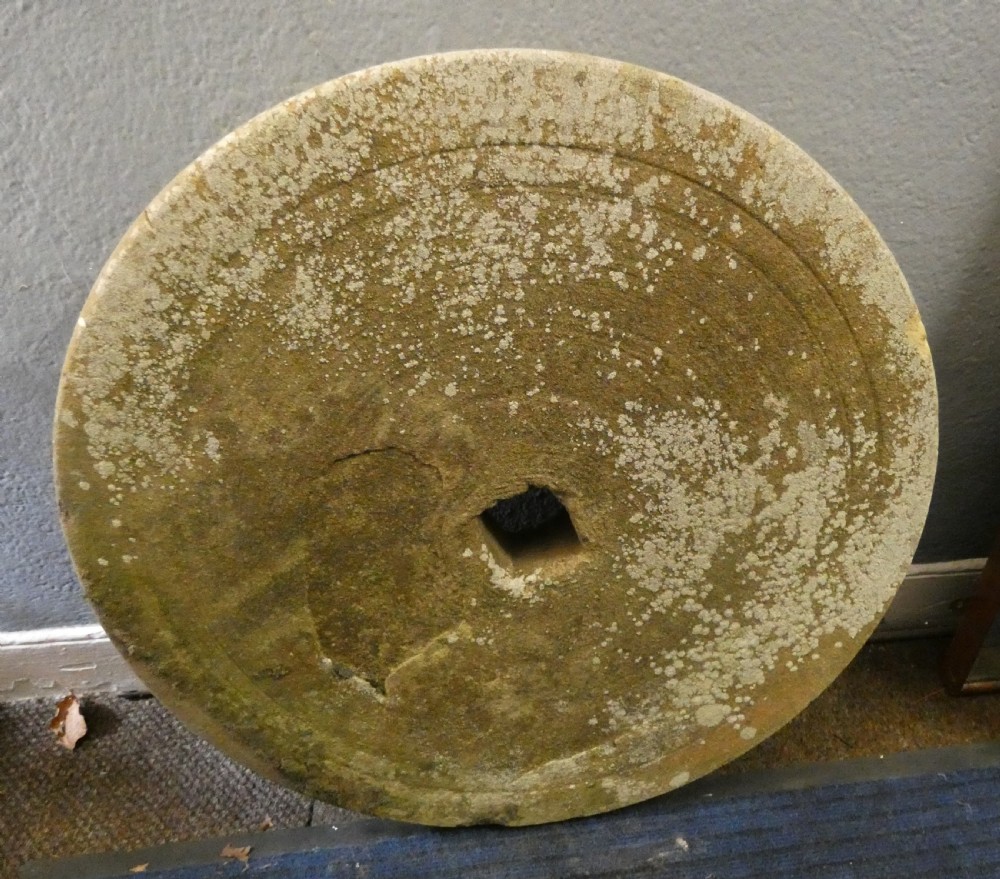 A circular hand-carved stone millstone featuring a square central aperture, concentric grooved circles, and a weathered moss-covered surface.